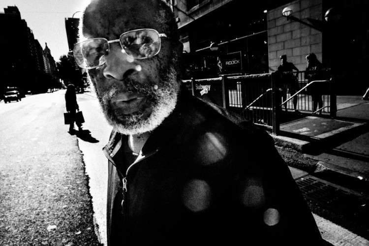 Black and white close photo of a bearded man in glasses looking into the camera on a New York sidewalk