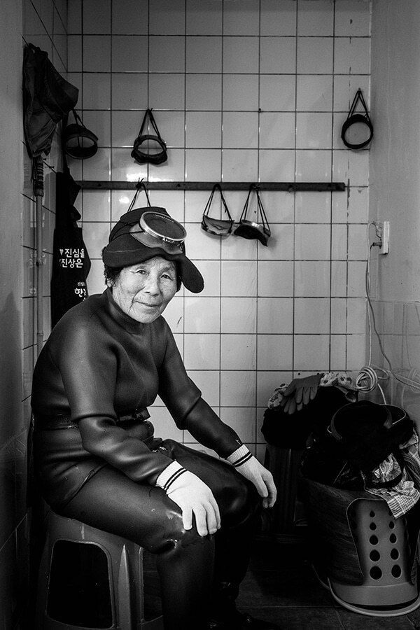 Environmental portrait of a Jeju haenyeo diver sitting on a stool in a tiled room with her diving gear hanging on the wall