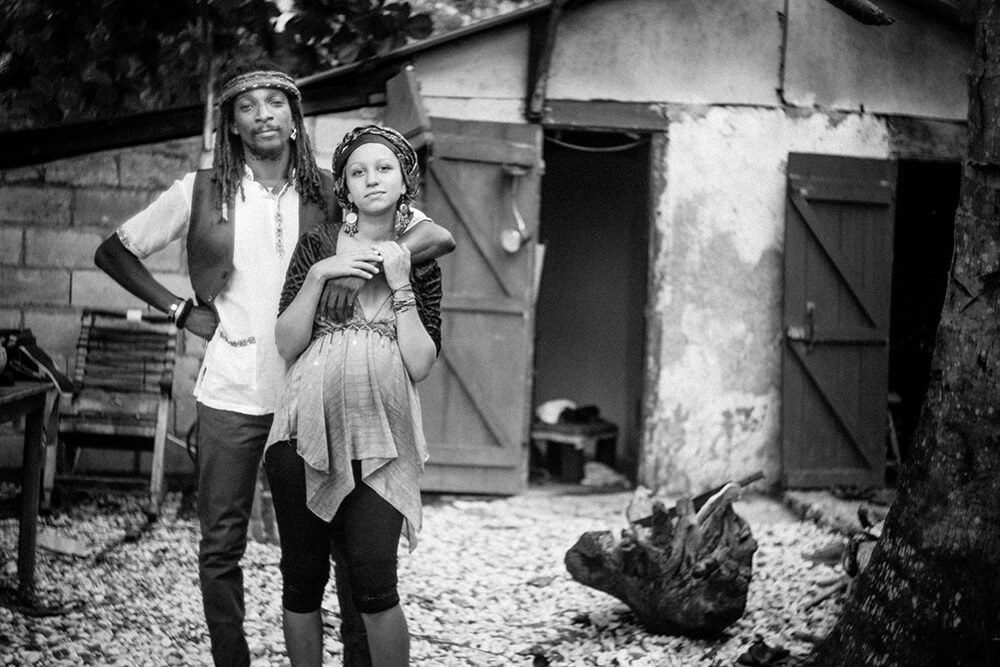 Black and white environmental portrait of a couple standing in front of a small wooden house, the man with his hand on his hip