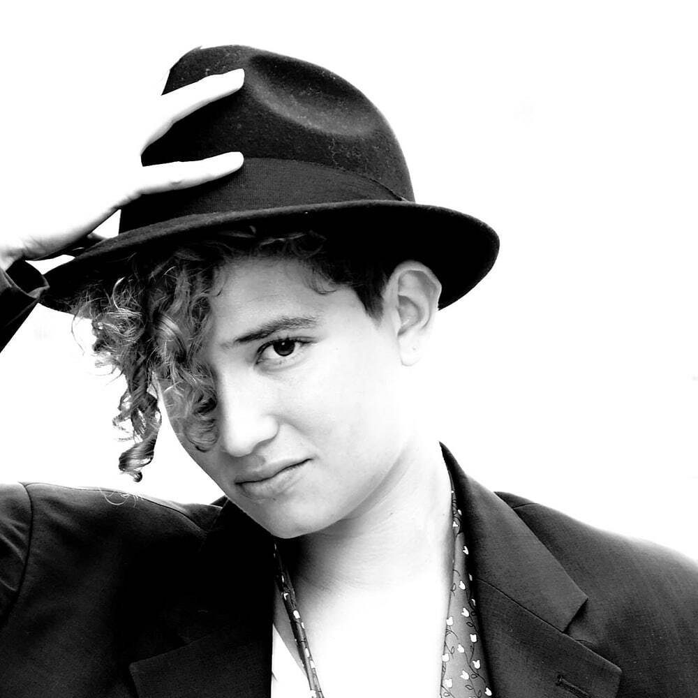 High key black and white portrait of a young woman holding the brim of her hat, with a white background