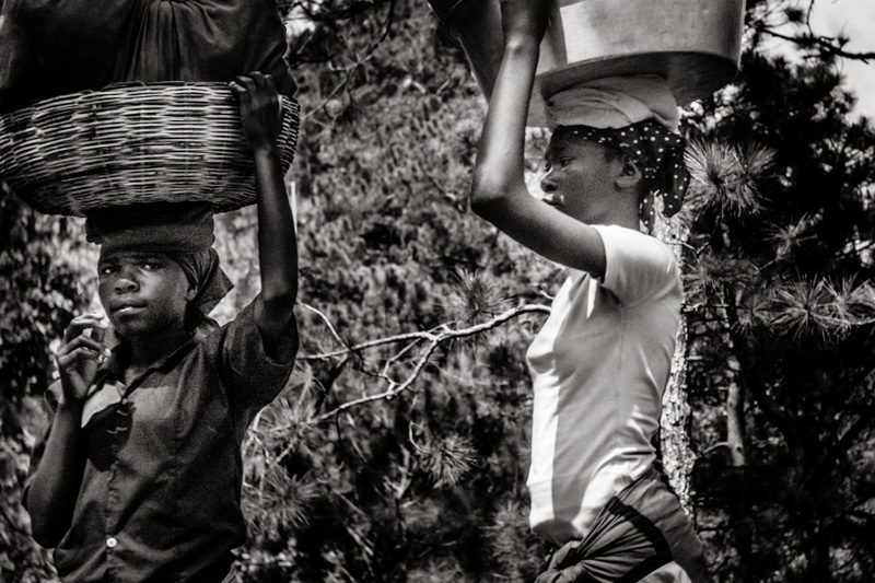 Black and white photo of a boy and a woman in Haiti carrying baskets on their heads under the trees