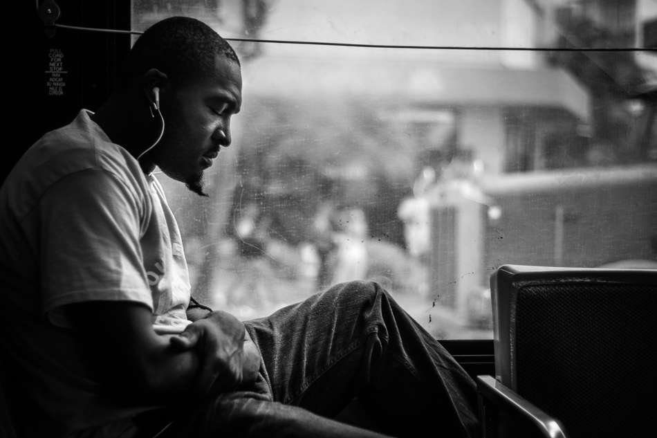 Black and white photo of a man with earphones sitting by a bus window, looking down, with a blurred street outside