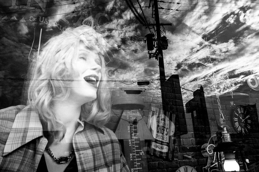Black and white reflection in a shop window mixing a laughing mannequin with city buildings and sky