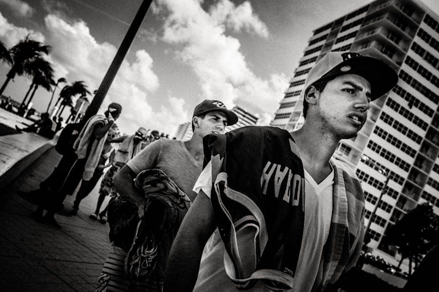 Low angle wide black and white street photo of young men walking past tall buildings under a cloudy sky