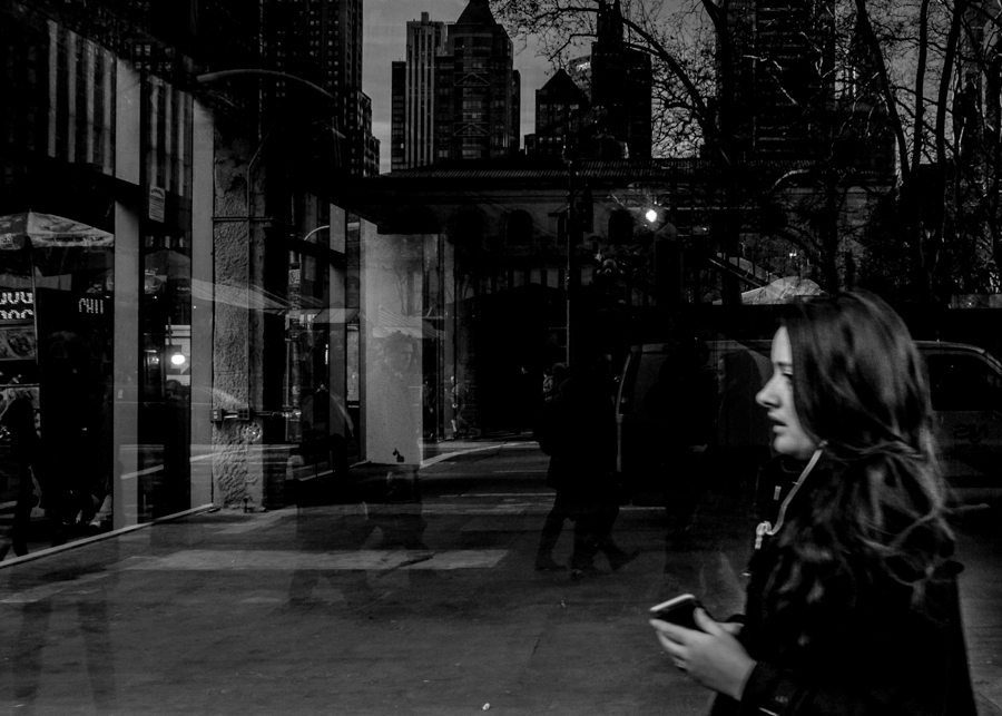 Black and white city street with a woman holding a phone in the foreground and buildings reflected in glass