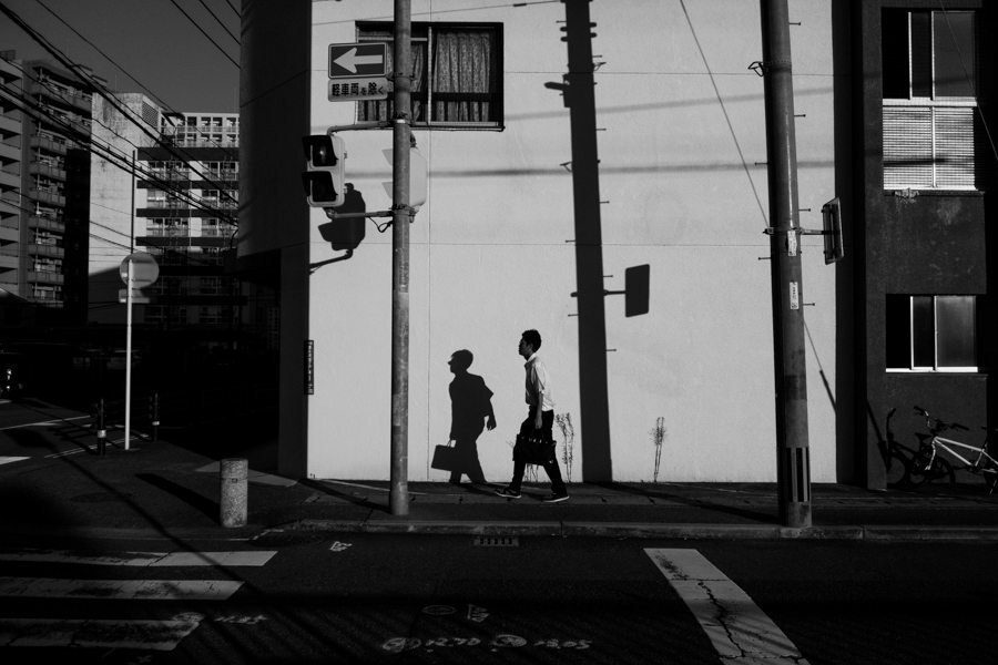 Black and white street scene with two pedestrians crossing between tall buildings and a utility pole