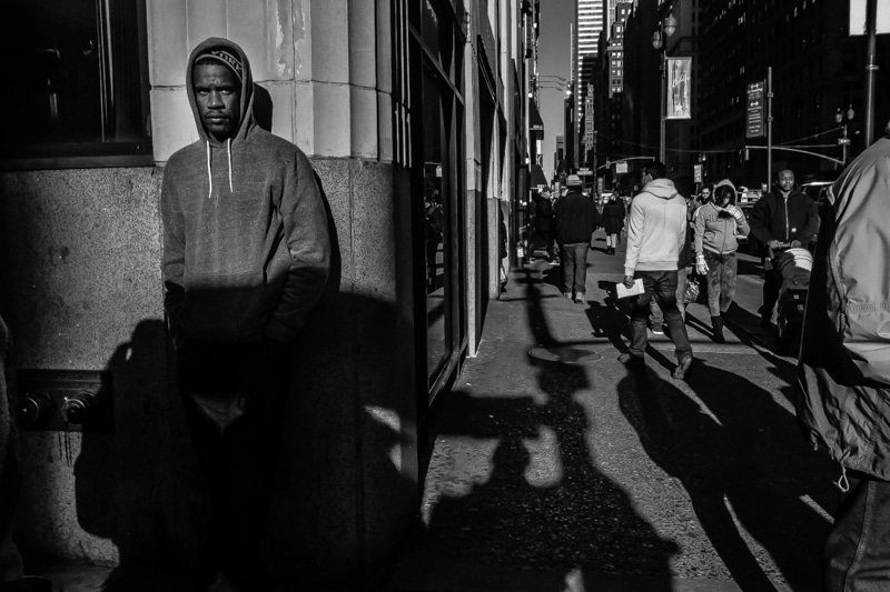 Man in a hooded jacket standing against a wall on a sunny sidewalk with pedestrians behind him