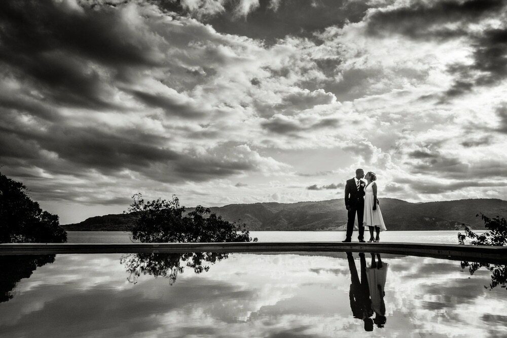 Couple standing at the edge of a still pool, reflected in the water under a dramatic cloudy sky in Haiti