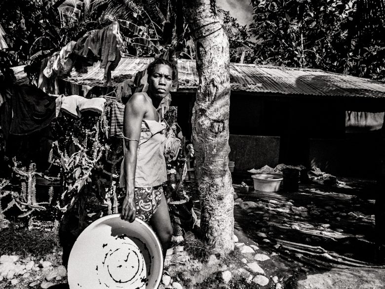 Black and white photo of a woman holding a plastic basin in a Haitian yard with laundry and trees behind her