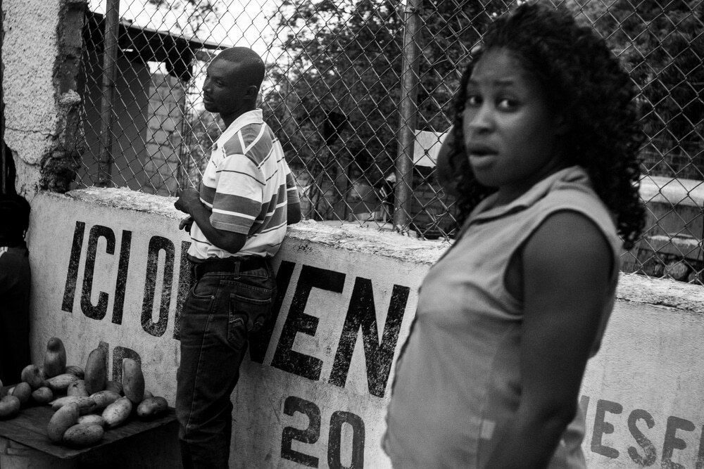 Black and white street photo of a woman looking at the camera beside a painted wall, a boy standing behind her
