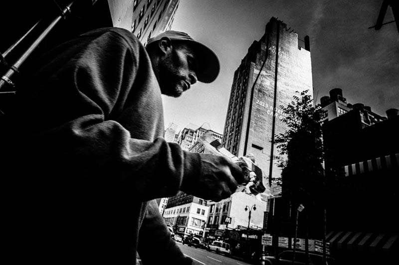 Low angle black and white photo of a man in a cap looking down, a tall building rising behind him