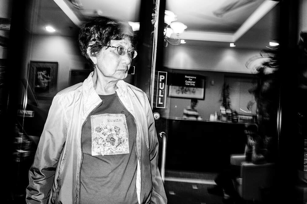 Black and white flash photo of an older woman in glasses walking through a shop