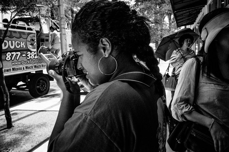 Black and white photo of a woman taking a picture with a camera on a busy city street
