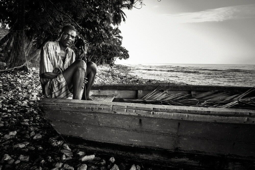Black and white photo of a man sitting in a wooden boat under a tree by the sea