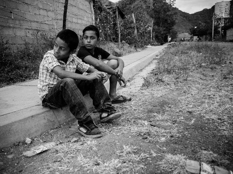 Black and white photo of two boys sitting on a curb beside a quiet road, taken up close