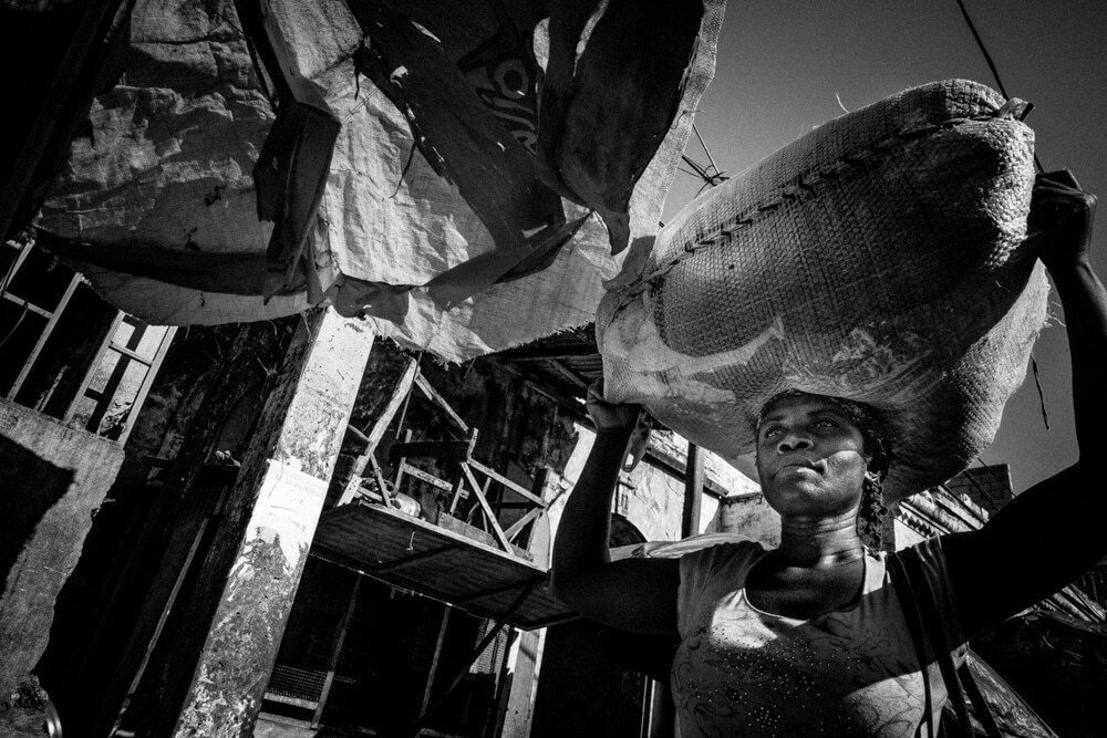 Low angle black and white photo of a woman carrying a large load on her head among market stalls