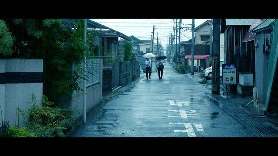 Wide color photo of a wet residential street in Japan on a rainy day, with two people walking away under a white and a black umbrella