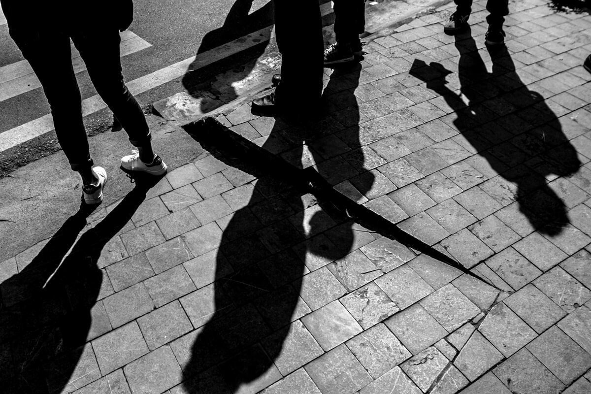 High contrast black and white street photo looking down at pavement with long hard shadows of passersby