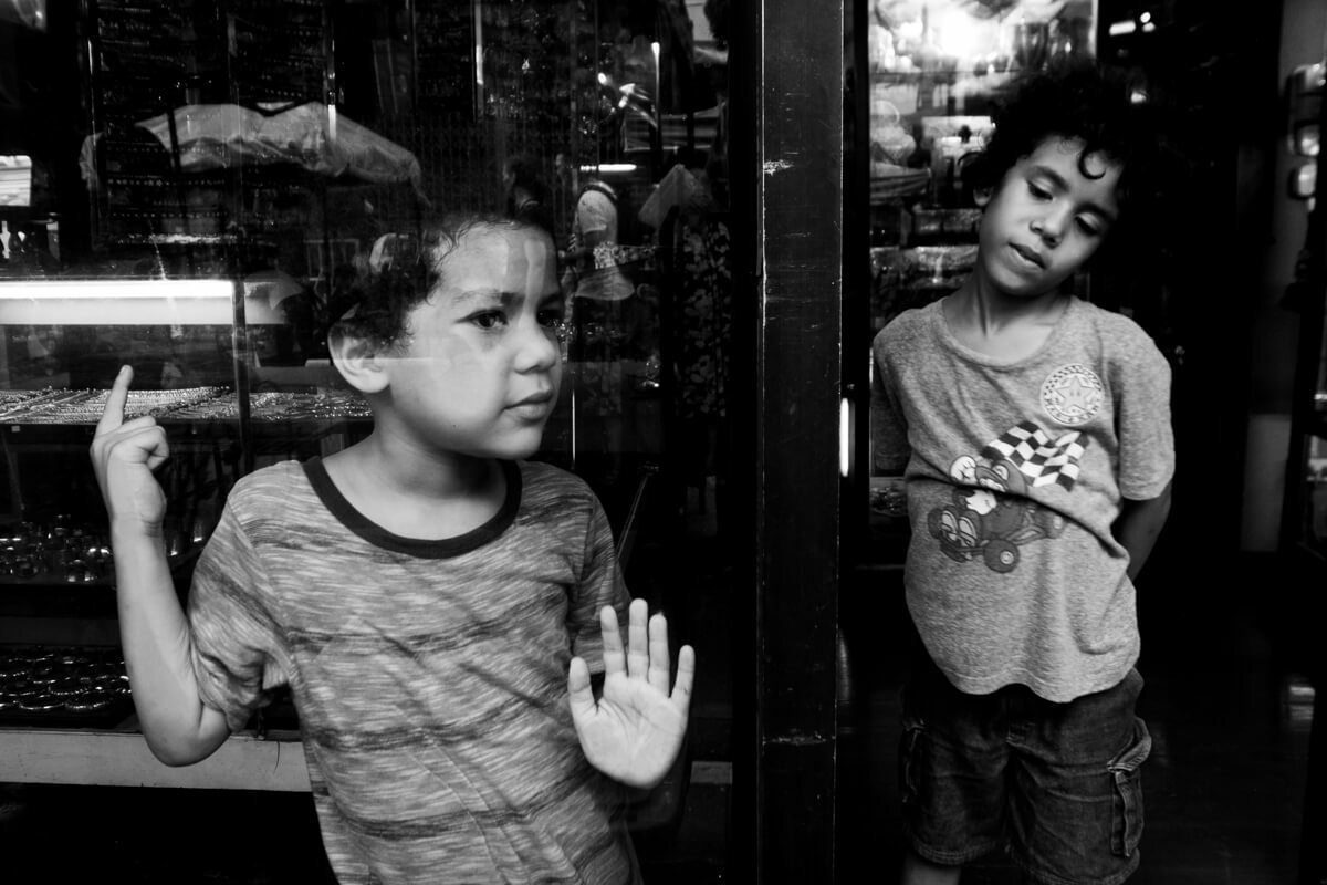 Black and white photo of two young boys behind a shop window, one raising his hand to the glass