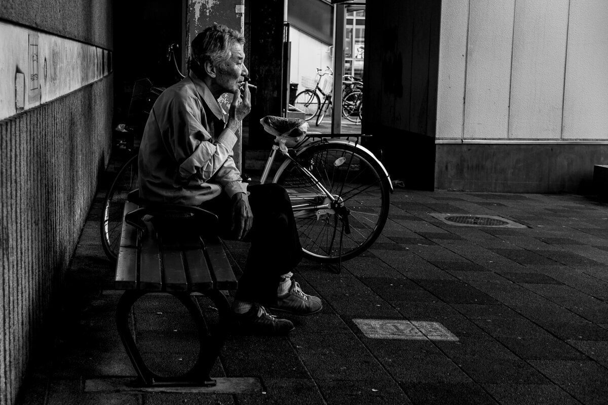 Black and white photo of an older man smoking on a bench next to a bicycle in a shadowy Japanese street
