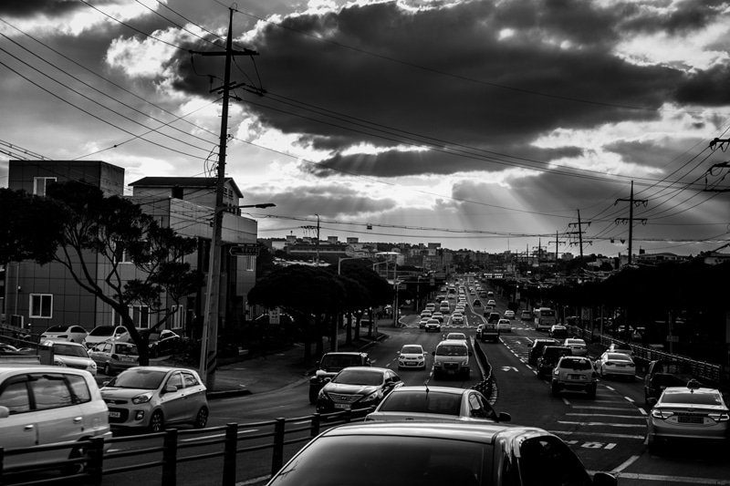 Black and white photo of a long road full of cars under dramatic clouds and power lines in Korea
