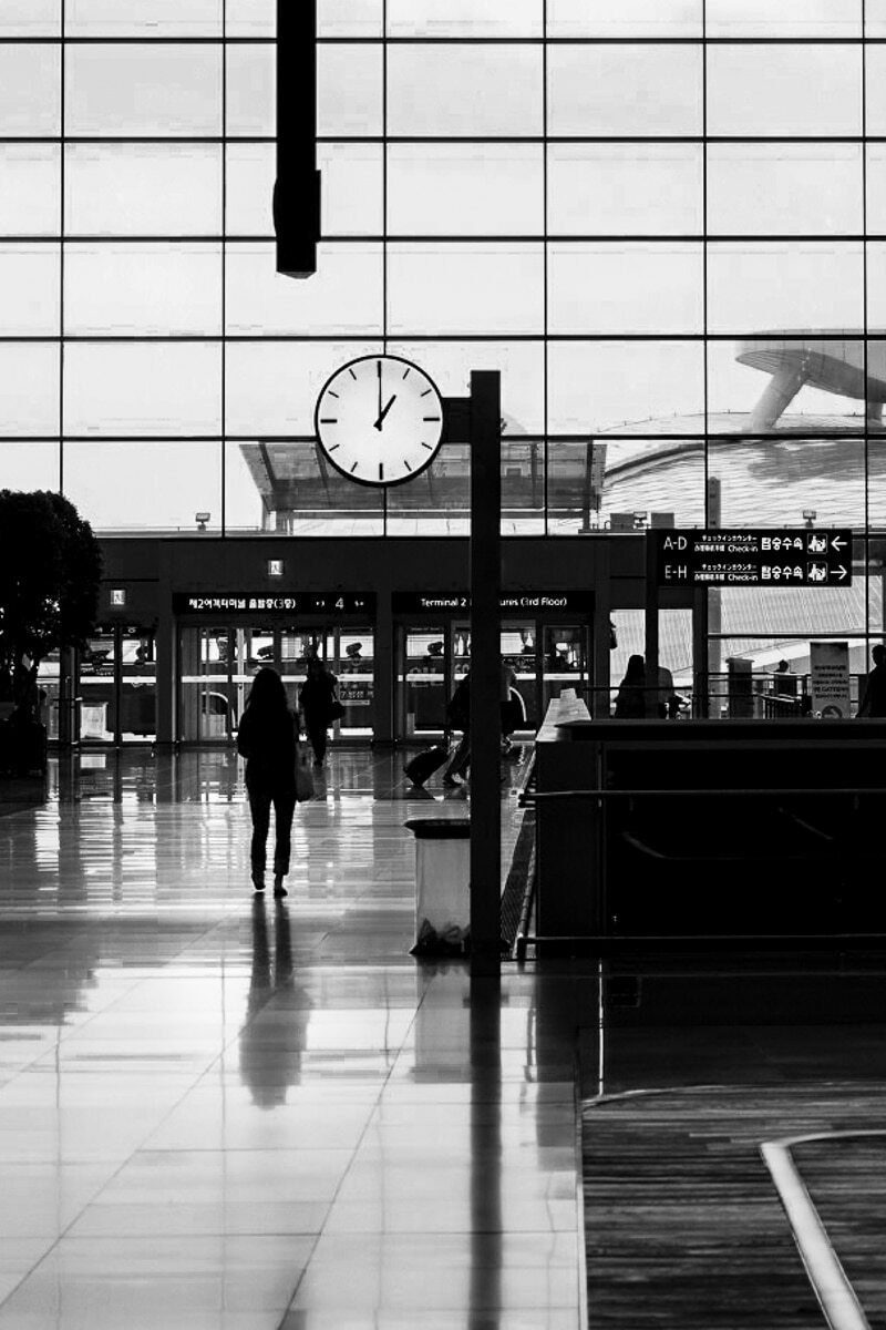 Black and white photo of a large airport hall with a single clock and a lone walking figure reflected on the shiny floor