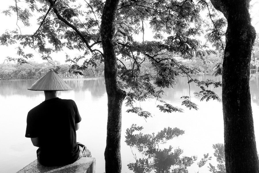 Black and white photo of a man in a conical hat sitting by a lake between two dark tree trunks