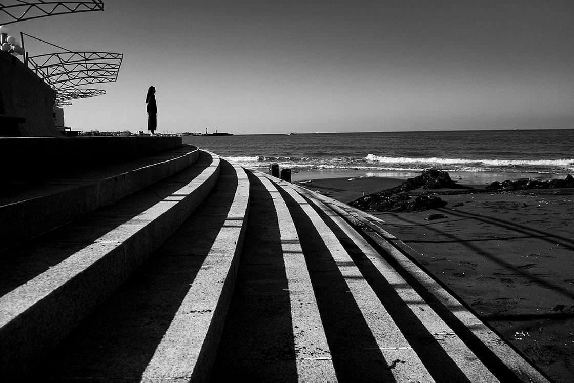 Black and white photo of curved concrete steps with repeating light and dark stripes leading to a lone figure standing by the sea