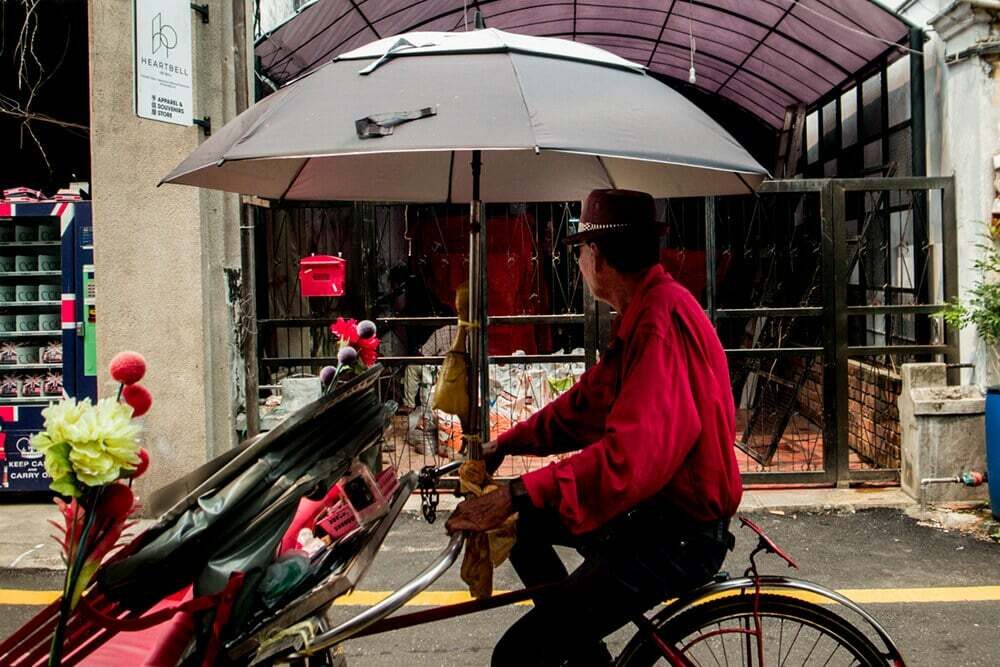 Man in a red shirt riding a trishaw decorated with red flowers under a large umbrella in Penang
