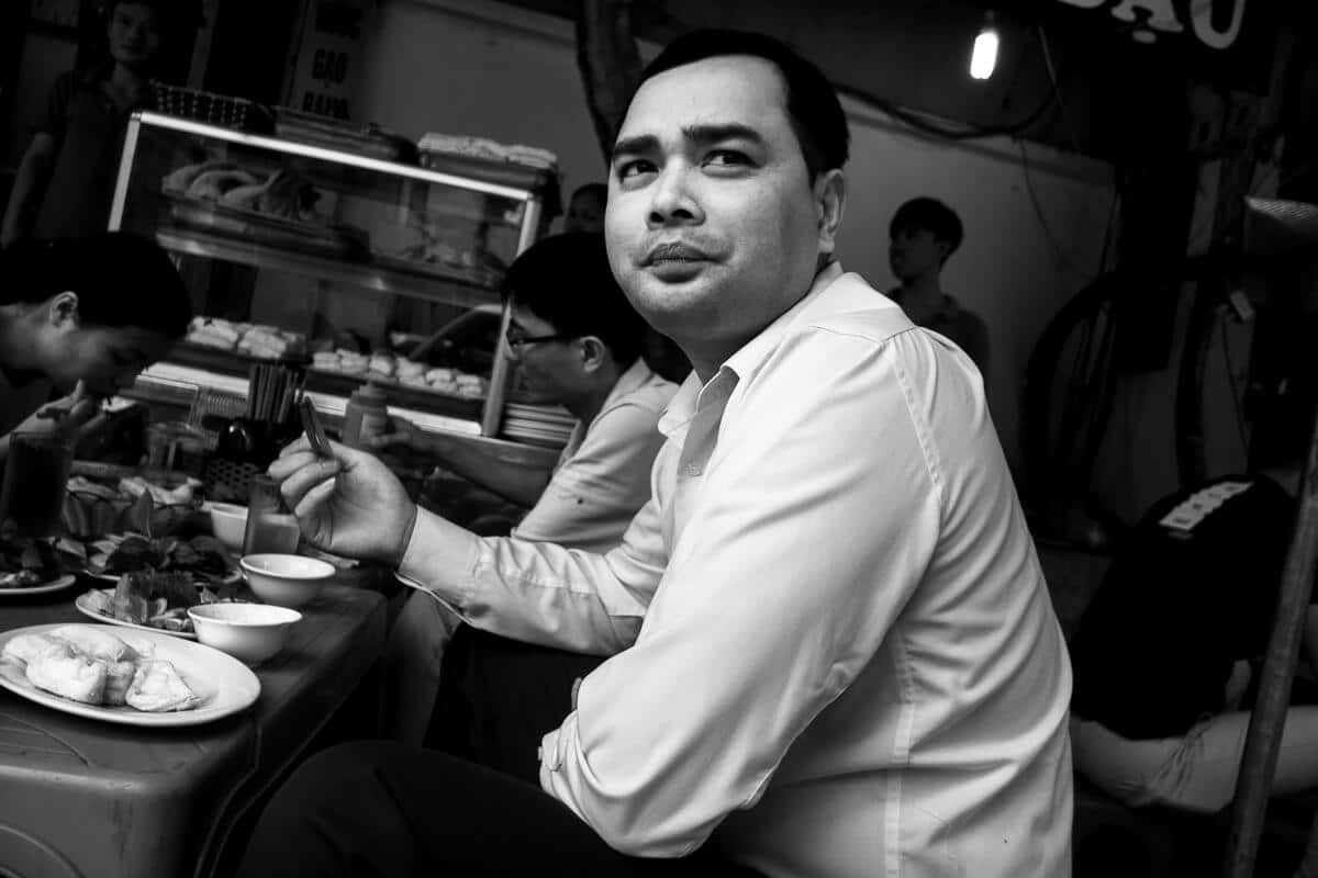 Man seated at a low street food table in Hanoi looking up at the camera
