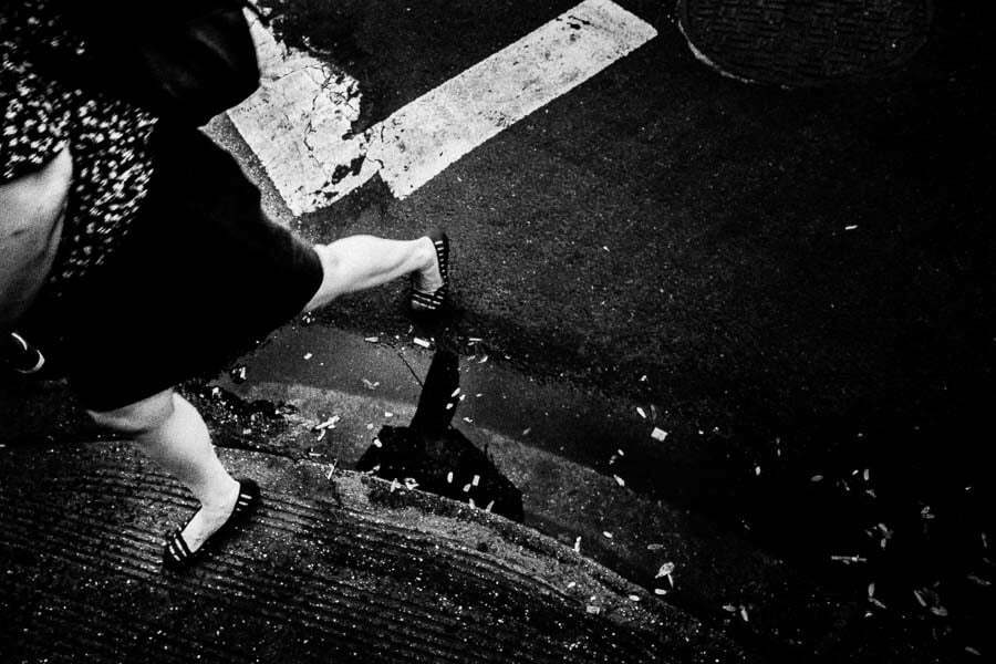 Black and white photo from a low angle of a woman's legs stepping off a curb onto a crosswalk stripe