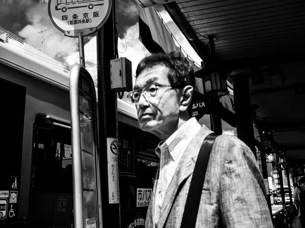 Black and white photo of a man in glasses at a Kyoto bus stop, framed by the bus stop sign pole