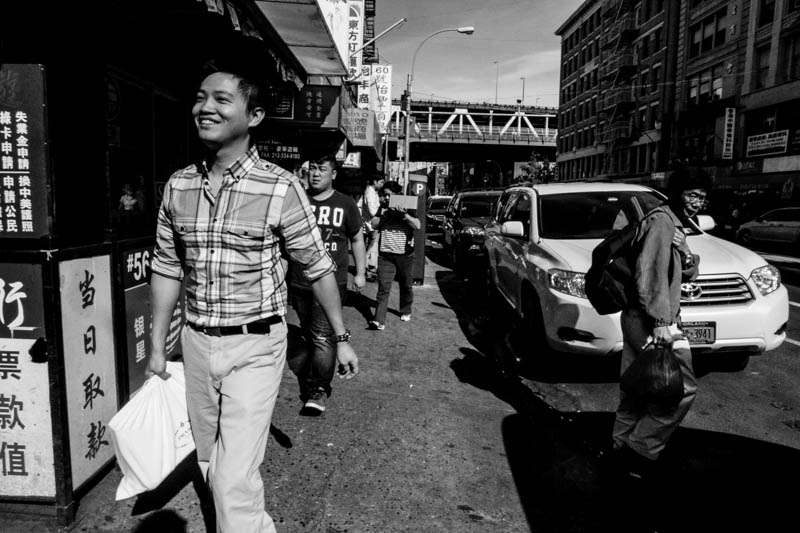 Black and white wide angle photo of a smiling young man in a checked shirt walking toward the camera on a busy Chinatown street