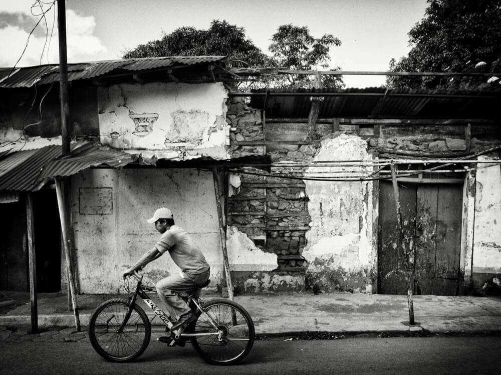 Black and white photo of a man riding a bicycle past a weathered wall with a tin roof in Nicaragua