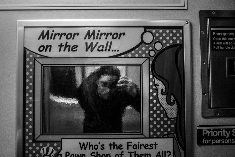 A pawn shop sign reading Mirror Mirror on the Wall, Who's the Fairest, with a blurred person seen through the glass inside the frame
