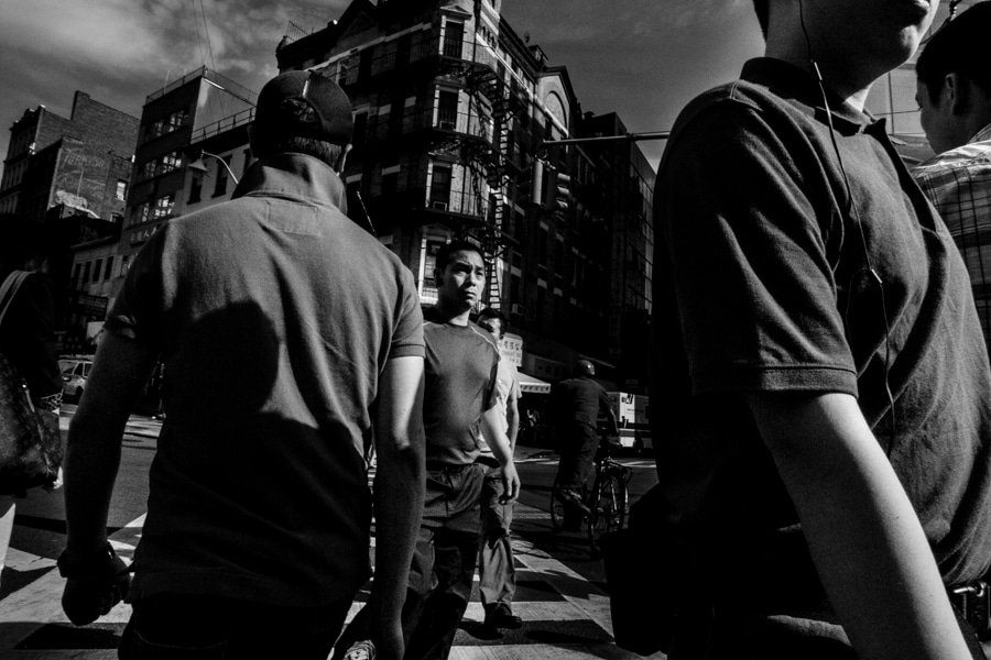 Black and white street scene shot from inside a crowd, with men walking close to the camera and a young man in the middle