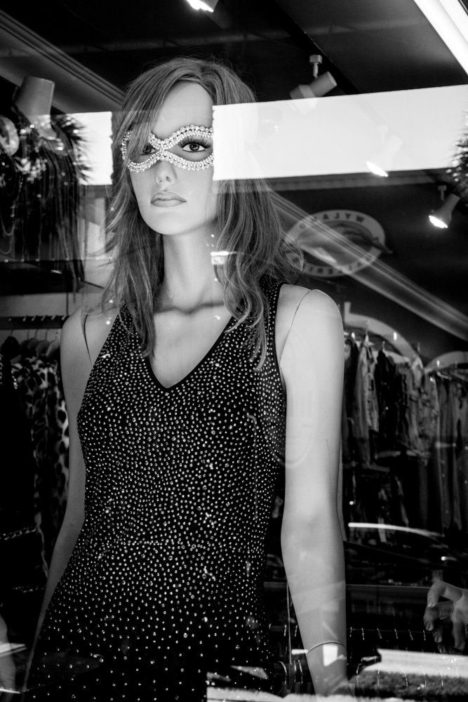 Black and white photo of a shop mannequin wearing a sparkly dress and a mask, seen through a window