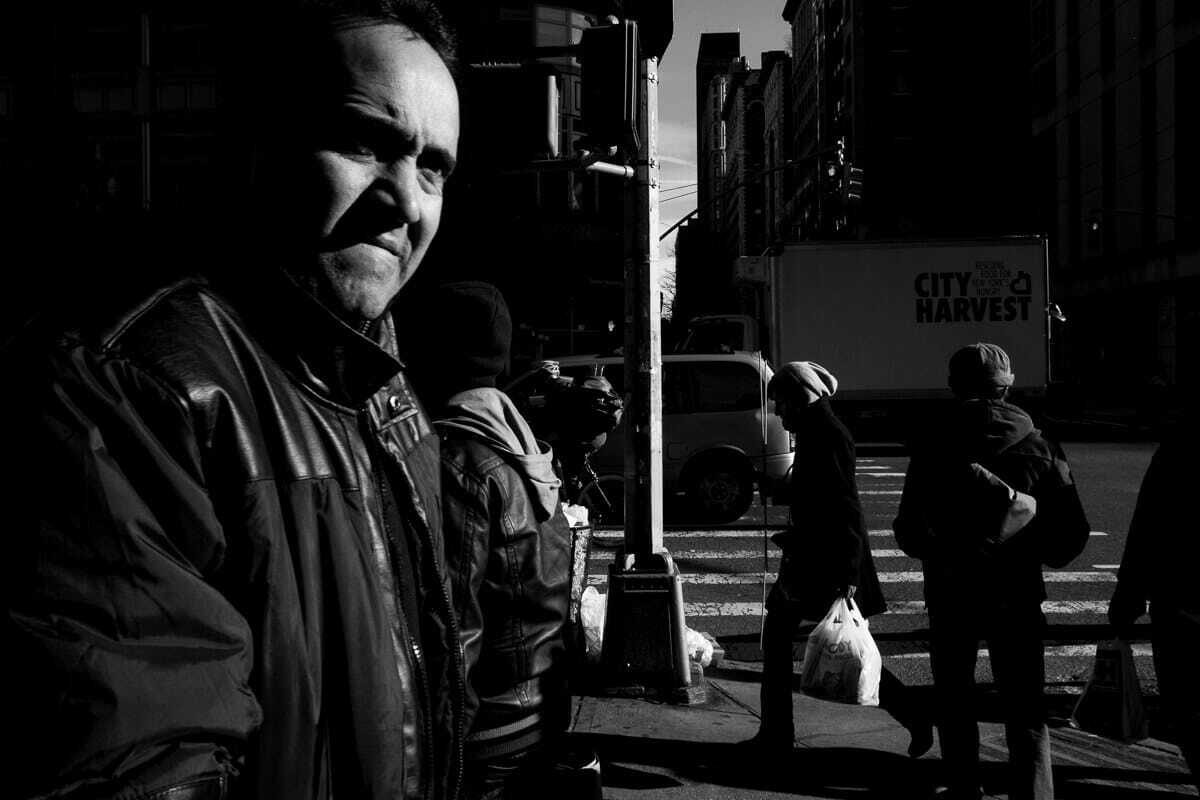 Black and white photo of a man in a leather jacket looking into the camera on a New York street corner