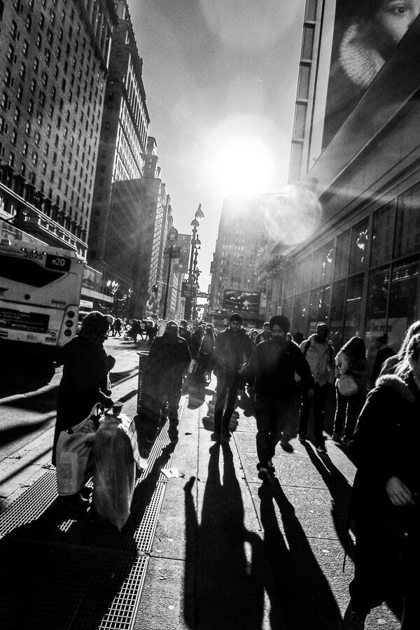 Vertical black and white photo of people walking on a sidewalk toward bright sun between tall buildings, long shadows behind them