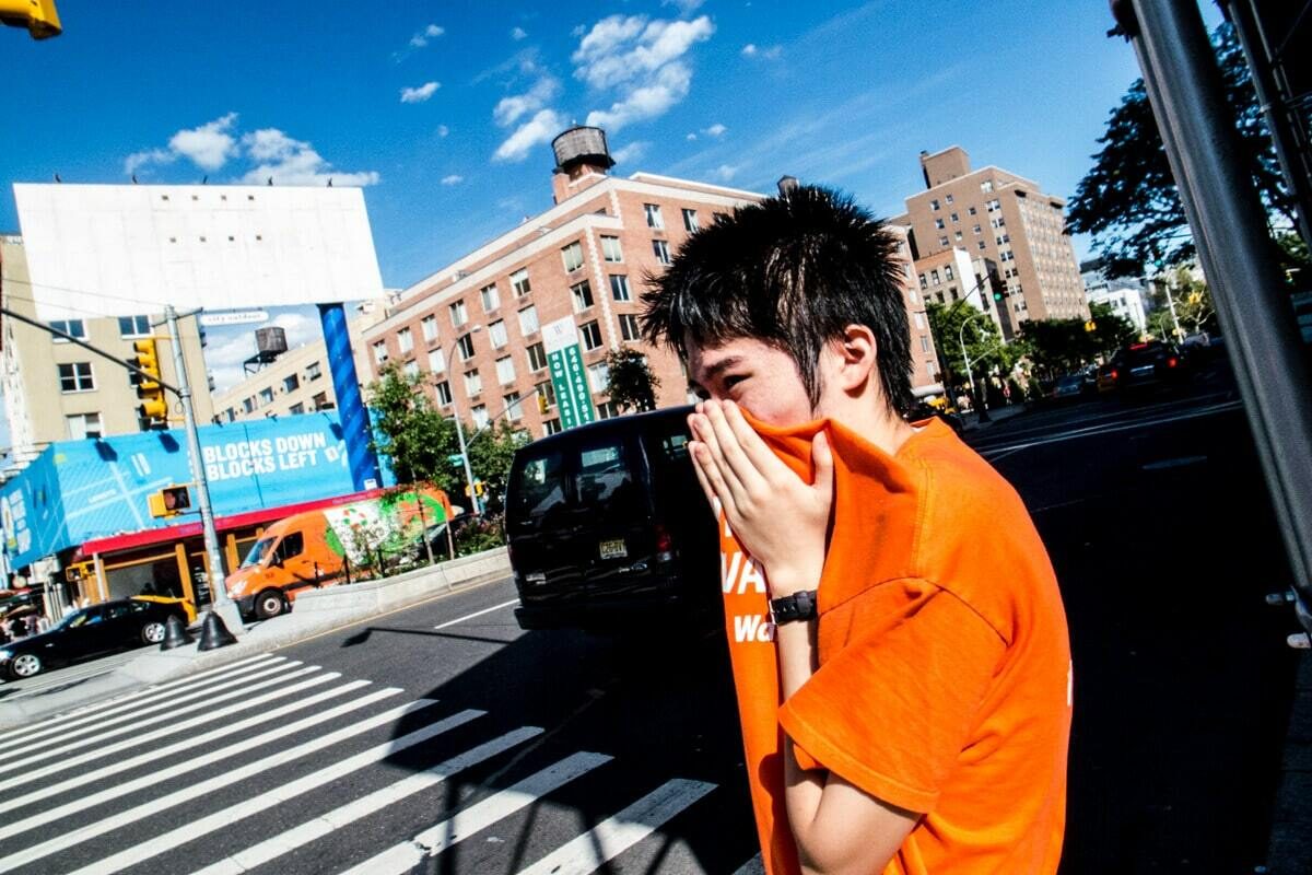 Colour photo of a young man in an orange shirt wiping his face, very close to the camera, at a street crossing