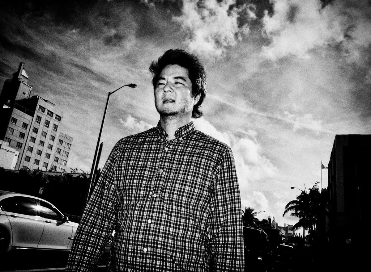 Low angle black and white photo of a man in a plaid shirt lit by hard sun against a cloudy sky