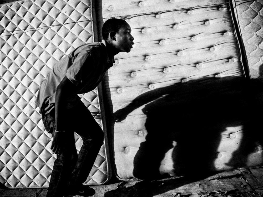 Black and white photo of a young man leaning past a mattress, with his large shadow cast across it