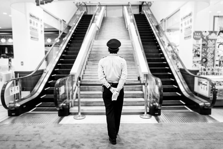 Black and white photo of a man in a uniform and cap walking toward a staircase between two escalators inside a mall in Japan