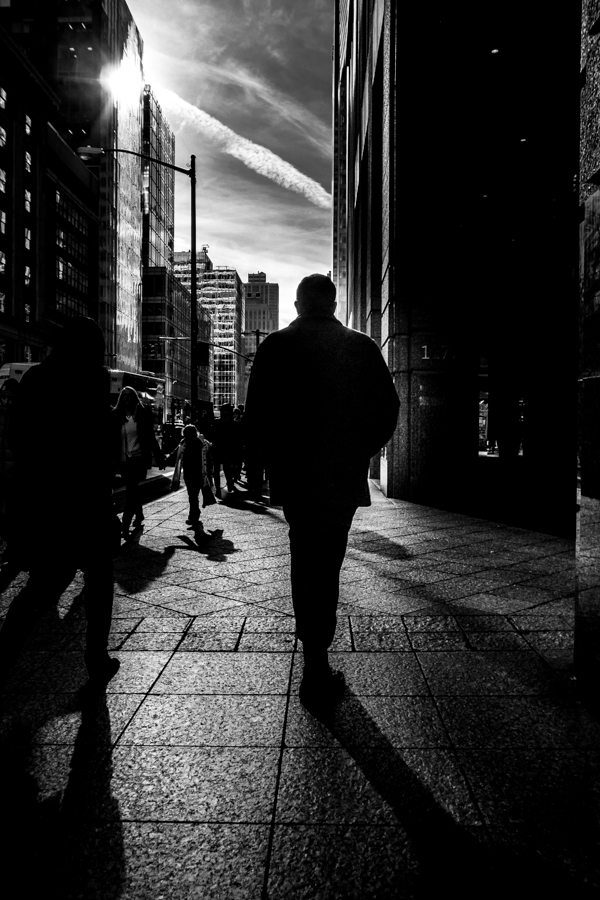 Black and white photo of a man walking on a city sidewalk into bright backlight, his shadow stretching toward the camera