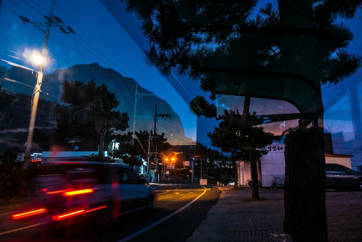 Street at dusk in Jeju, Korea, with a blue sky over a mountain, a street lamp and red car light trails