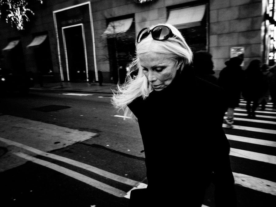 Black and white street photo of a woman with sunglasses on her head crossing a street, looking down