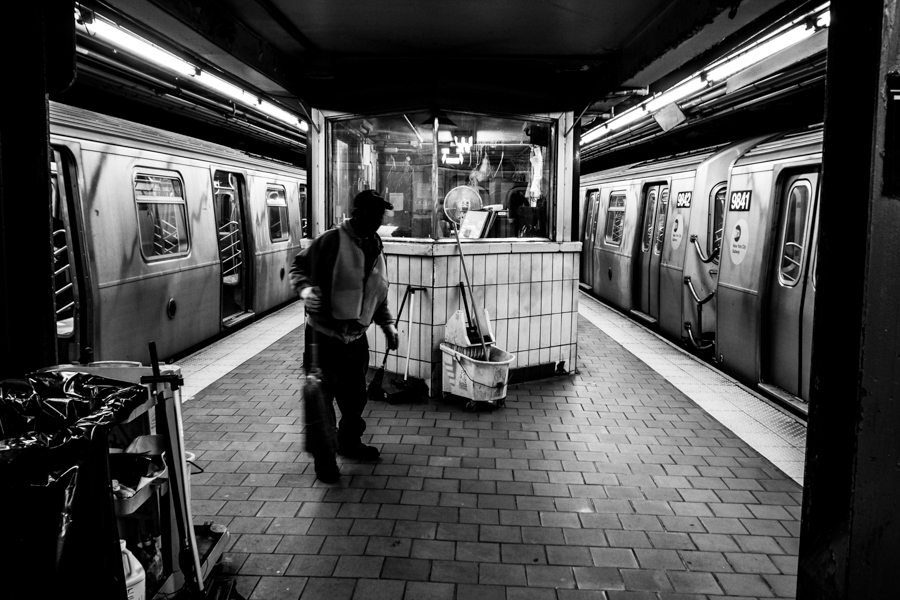 Black and white photo of a subway platform with a worker pushing a cart between two trains
