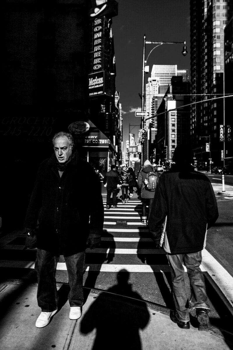 Vertical black and white street photo of two men walking toward the camera on a crosswalk between tall buildings