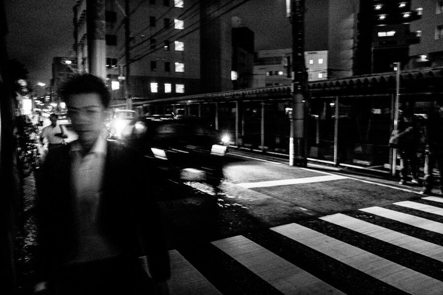 Black and white night photo of a man in a white shirt blurred as he walks past a crosswalk