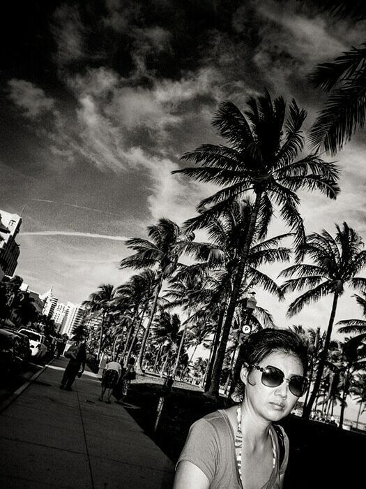 Black and white photo of a woman in sunglasses in front of tall palm trees under a bright sky